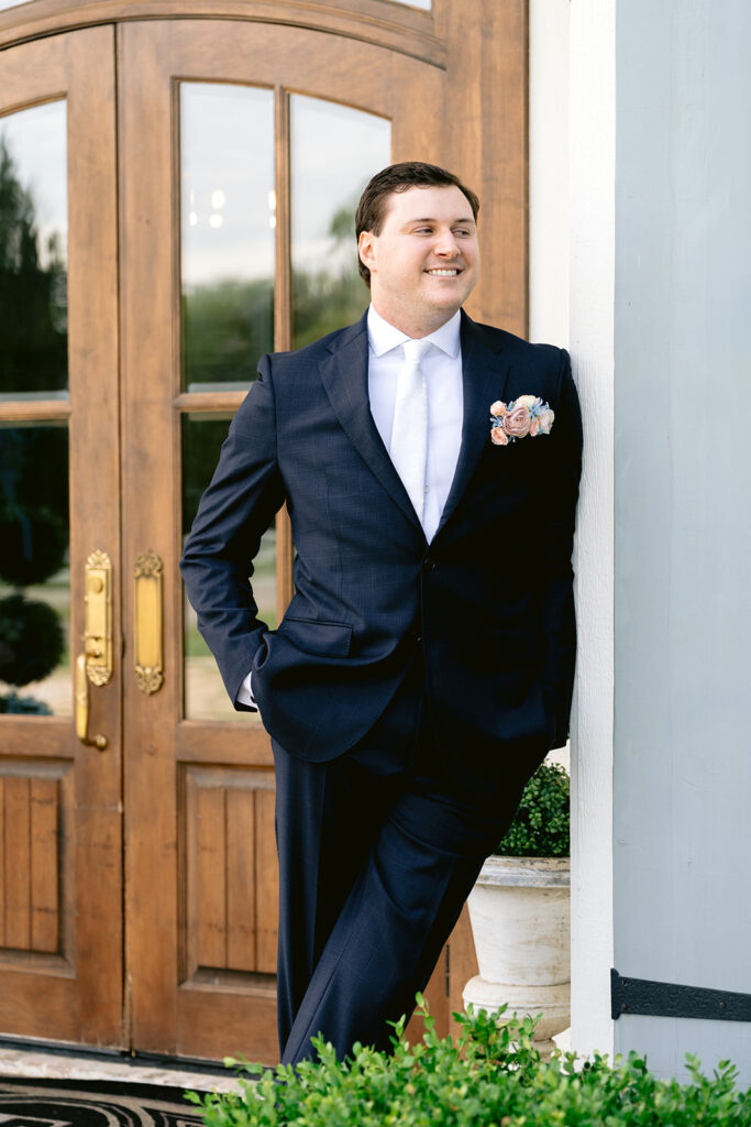 Classic groom portraits showcasing the groom in a tailored black suit with a blush floral boutonniere, captured outside The French Farmhouse wedding venue in Collinsville, Texas before the wedding ceremony.